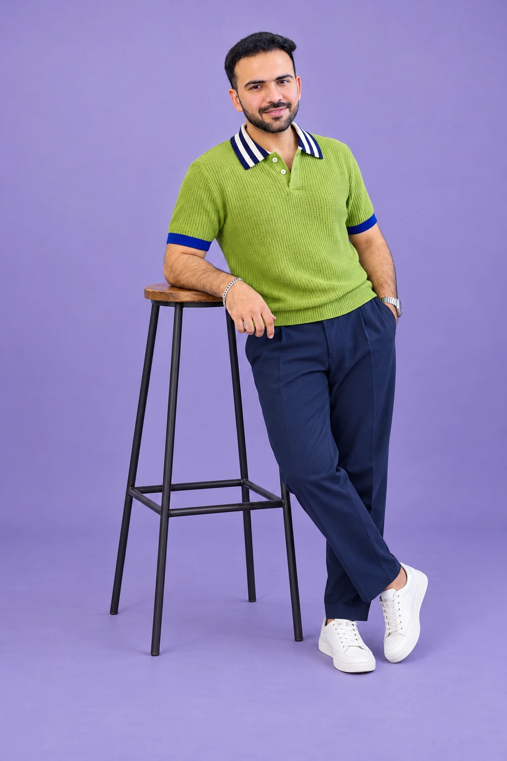 Full-length portrait of Shivam in a lime knit polo beside a wooden stool